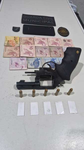 Imagem de uma arma de fogo, munições, dinheiro em cédulas e identificação da Guarda Civil Municipal dispostos sobre uma mesa.