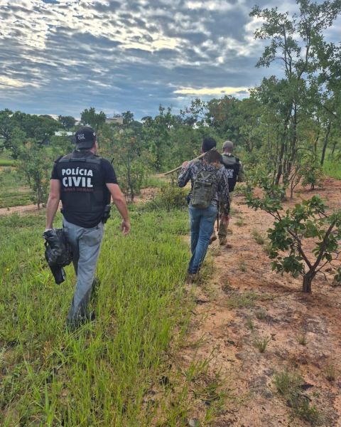 Agentes da Polícia Civil caminhando em uma área rural, cercados por árvores e vegetação, enquanto observam a paisagem.