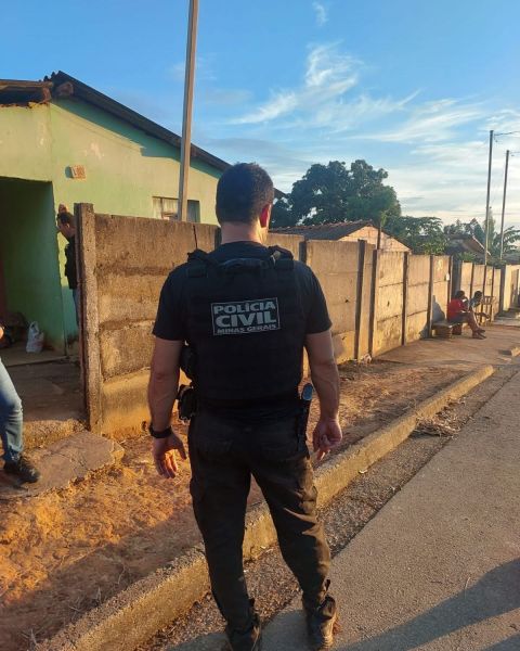 Um policial civil de costas em uma rua, com a identificação 'Polícia Civil' visível em seu colete, perto de casas e pessoas nas proximidades.