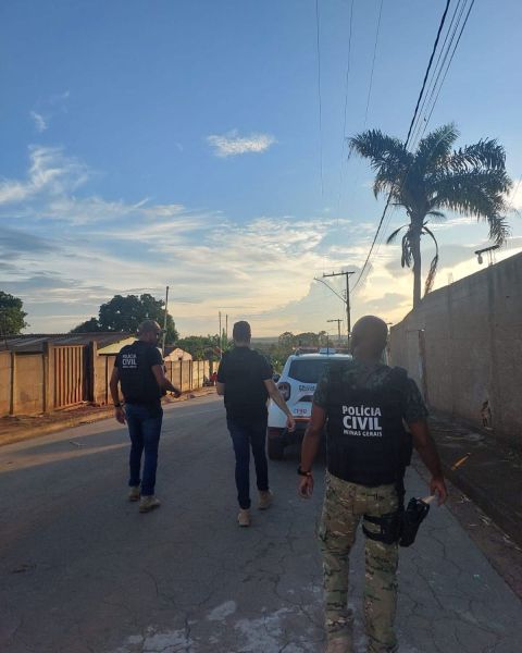 Policiais civis caminhando em uma rua de bairro, com um veículo da polícia ao fundo sob um céu claro ao entardecer.