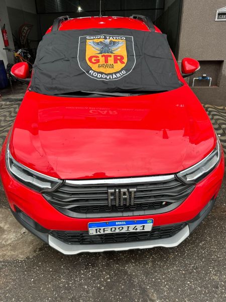 Uma visão frontal de um carro vermelho da marca Fiat, com uma toalha de proteção preta com um escudo amarelo e vermelho do Grupo Tático Rodoviário cobrindo o para-brisa.