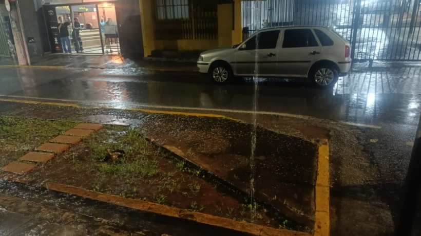 Imagem de uma rua molhada à noite, com um carro estacionado na calçada e água escorrendo de um bueiro.