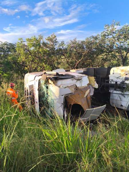 Caminhão acidentado deitado na estrada, com danos visíveis na carroceria, enquanto um socorrista em roupa laranja trabalha nas proximidades cercado por vegetação alta.