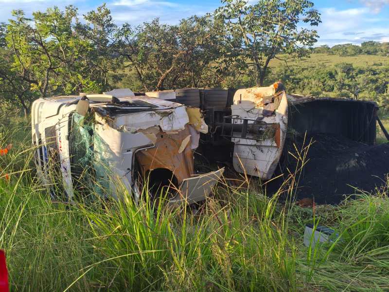Caminhão acidentado tombado em uma área rural, com danos visíveis na cabine e vegetação ao redor.