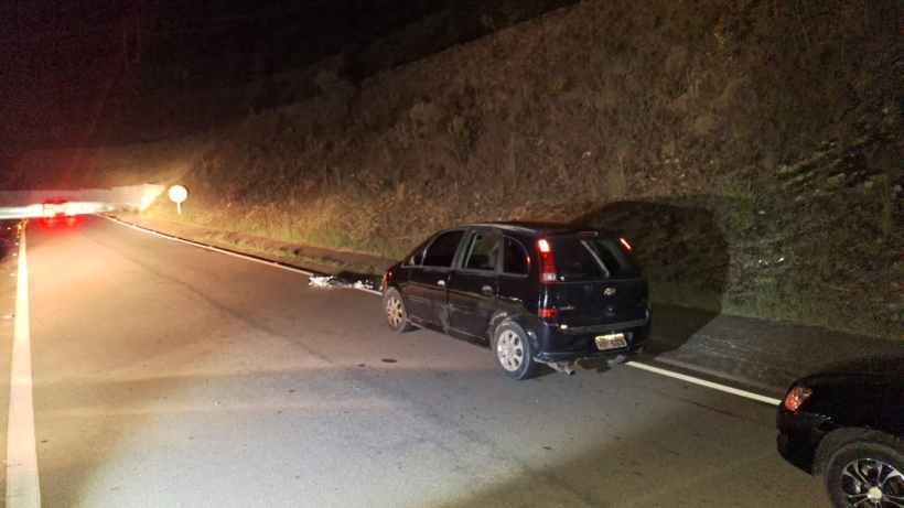 Imagem de um carro preto parado na beira de uma estrada à noite, com sinalização visível e vegetação ao fundo.