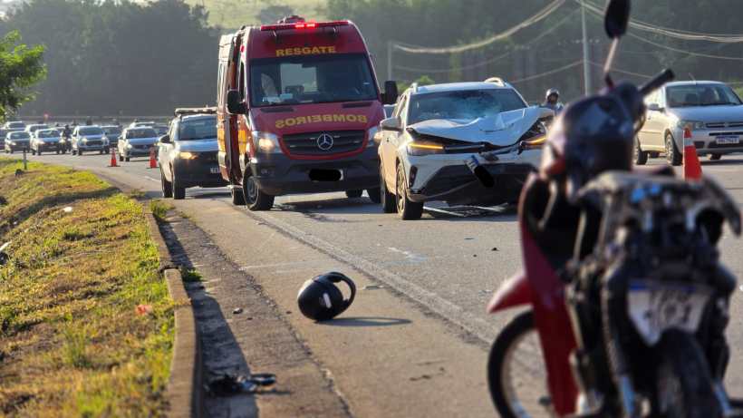 Veículo de resgate ao lado de um carro colidido em uma estrada, com tráfego parado ao fundo e um capacete caído no chão.