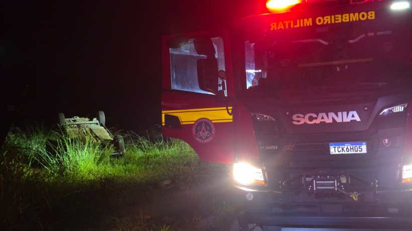 Caminhão dos bombeiros iluminado à noite com um veículo tombado ao fundo em uma área com grama alta.
