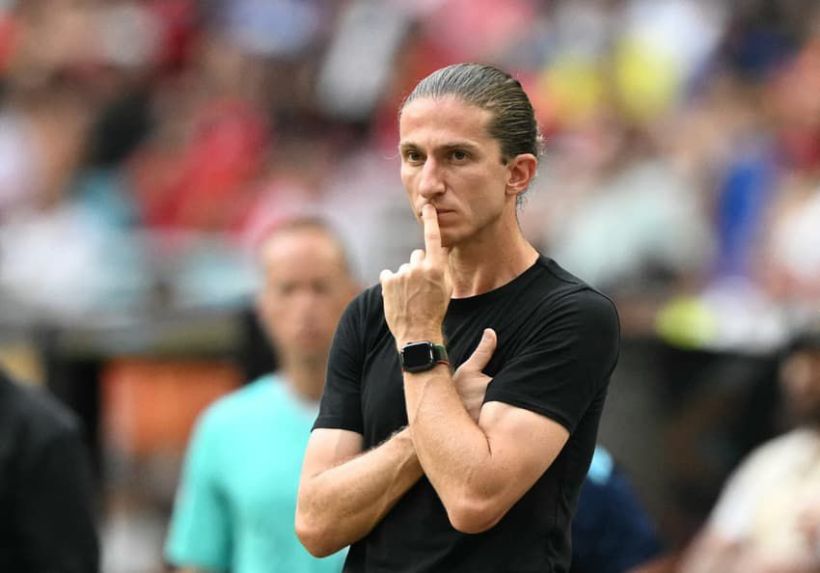 A serious-looking coach with long hair pulled back is standing on the sidelines, thoughtfully resting a finger on his lips, as he observes the game.
