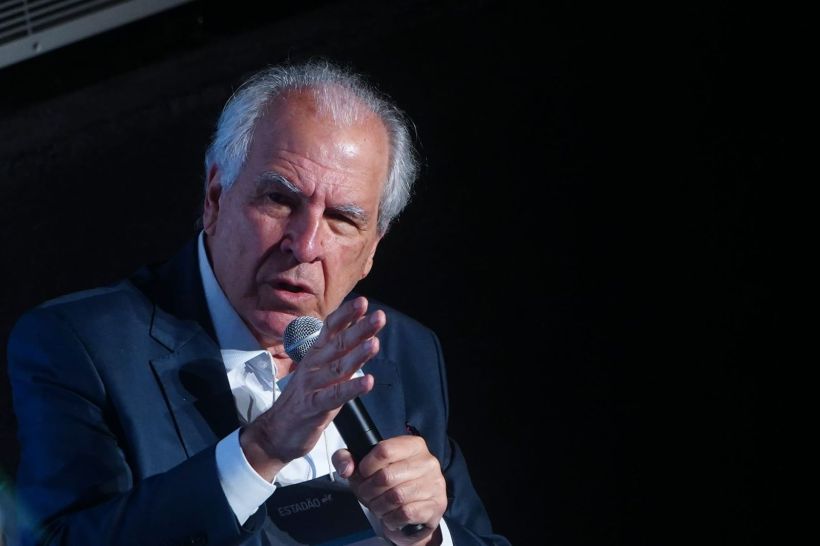 A man in a dark suit passionately speaking into a microphone, gesturing with his hands against a dark background.