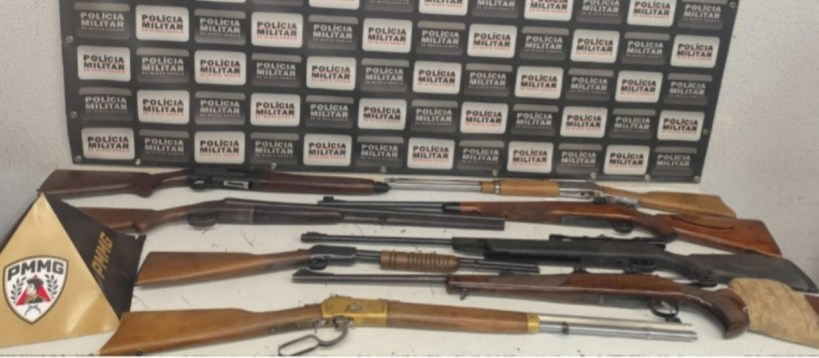 Armas de fogo expostas em uma mesa, com um painel da Polícia Militar ao fundo.