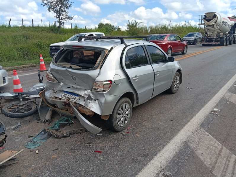 Carro prateado danificado em acidente, com a traseira amassada, ao lado de uma motocicleta caída na estrada.