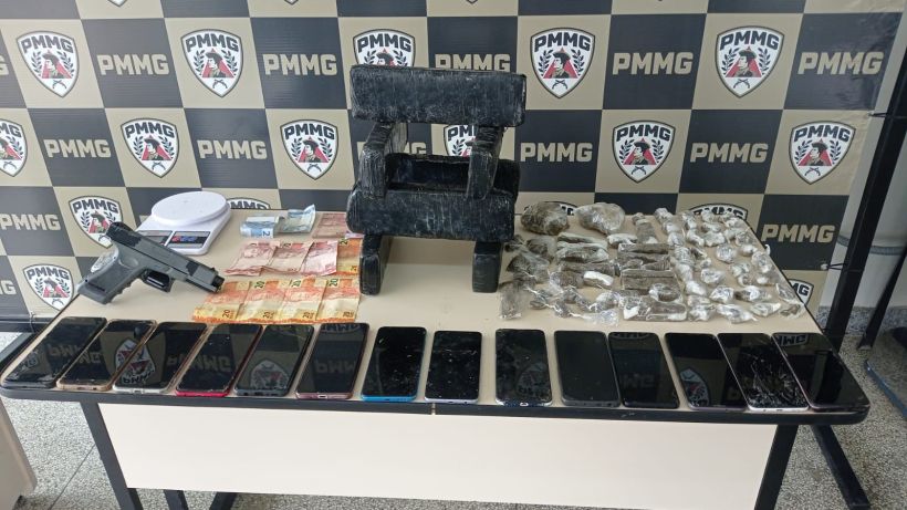 Mesa com diversos items, incluindo uma arma de fogo, dinheiro em cédulas, uma balança, um pedaço de concreto, e vários celulares dispostos em fileira, em frente a um fundo com o logo da PMMG.