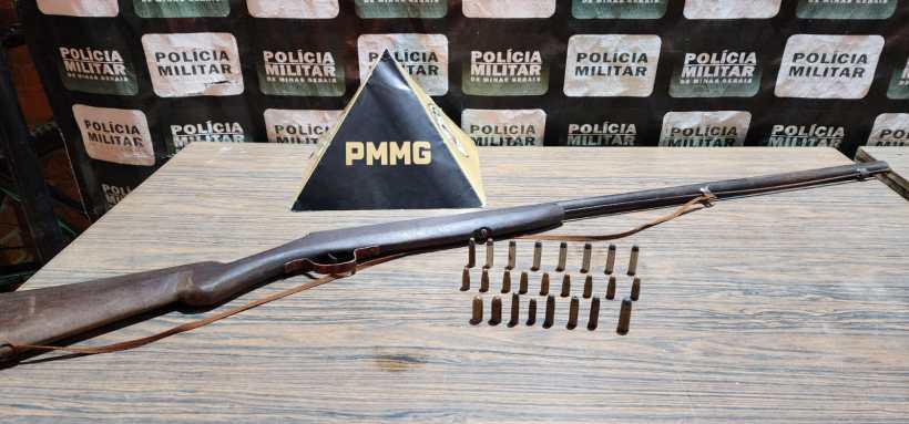 Rifle antigo ao lado de cartuchos de munição em uma superfície de madeira, com fondu de fundo da Polícia Militar.