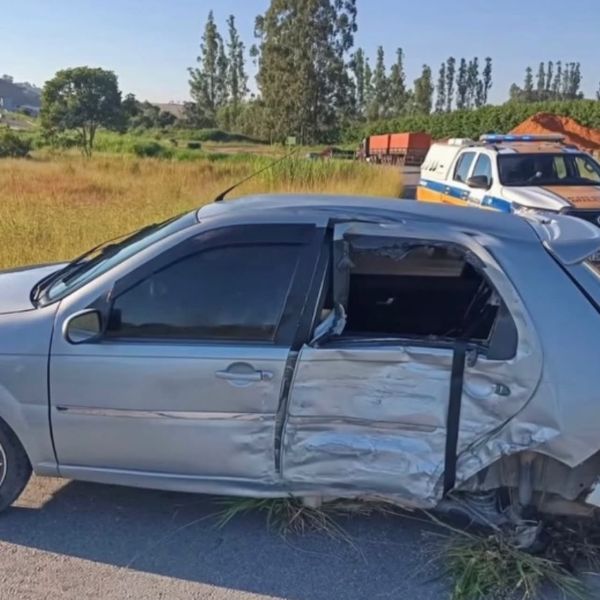 Carro prata danificado com a porta aberta em uma área rural, ao fundo há um veículo de polícia e vegetação ao redor.