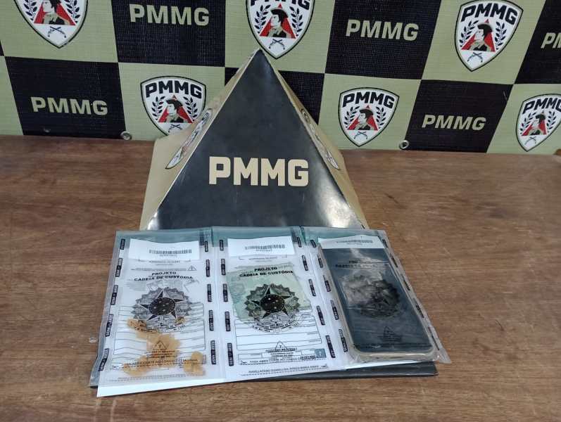 Um suporte em forma de pirâmide com a inscrição PMMG, com documentos e um distintivo sobre uma mesa, em frente a um fundo com logotipos da PMMG.