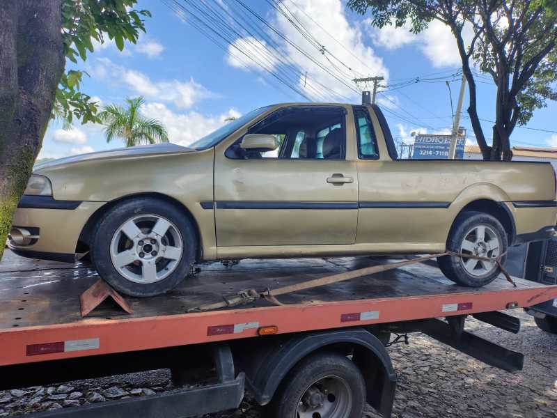 Um carro pick-up de cor dourada carregado em um caminhão guincho, estacionado sob árvores e com um céu claro ao fundo.