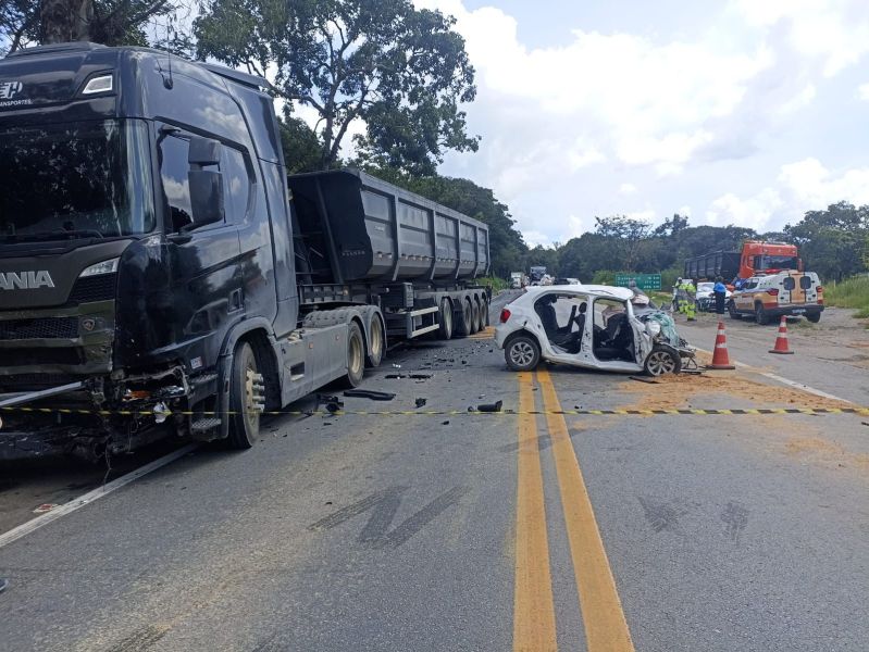 Acidente de trânsito envolvendo um caminhão e um carro de passeio em uma estrada. O carro está danificado e inclinado, enquanto o caminhão está estacionado ao lado. Há cones de sinalização e equipes de emergência na cena.