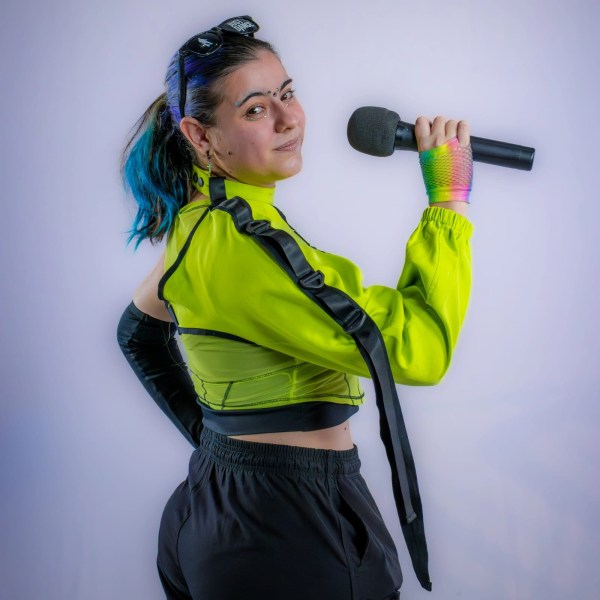 Pessoa com cabelo colorido e blusa fluorescente, segurando um microfone e sorrindo, em um fundo claro.