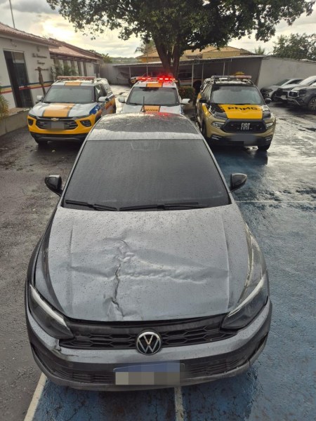 Um carro Volkswagen danificado, com a frente amassada, estacionado em frente a viaturas de polícia com luzes de emergência ligadas.
