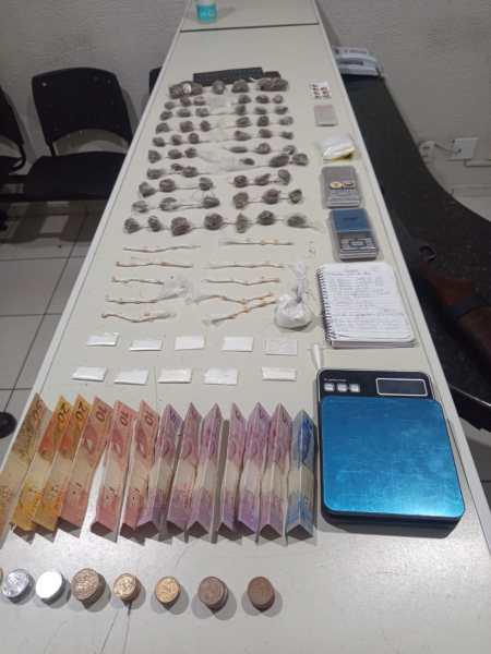 Mesa com diversos itens relacionados a tráfico de drogas, incluindo pacotes, balança, dinheiro em notas e moedas, além de uma agenda.