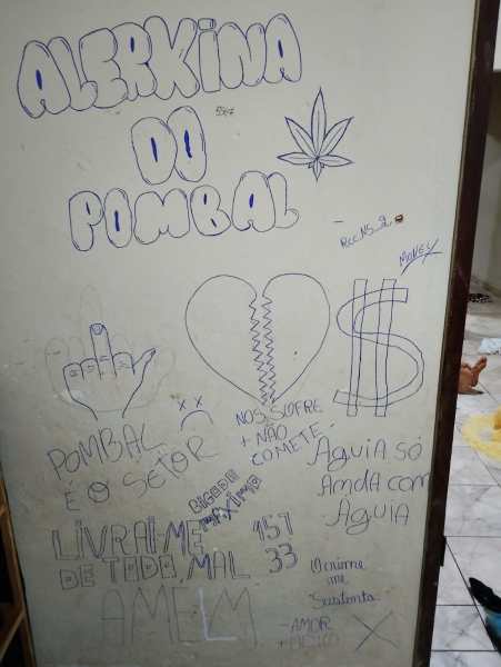 Parede com graffiti, incluindo frases e desenhos como um coração quebrado, um símbolo de dólar, uma folha de maconha e gestos de mãos.