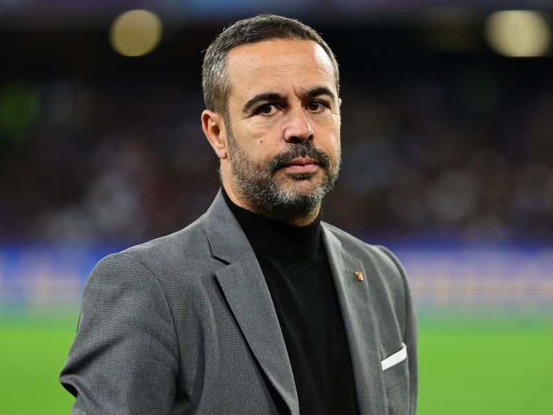 A man with short hair and a beard, wearing a gray blazer over a black turtleneck, standing on a football field with a focused expression.