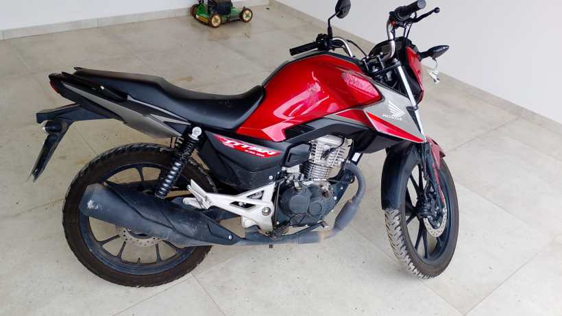 Uma motocicleta Honda CG 160 Titan em destaque, com carroceria vermelha e detalhes pretos, estacionada em uma área interna com piso de cerâmica.