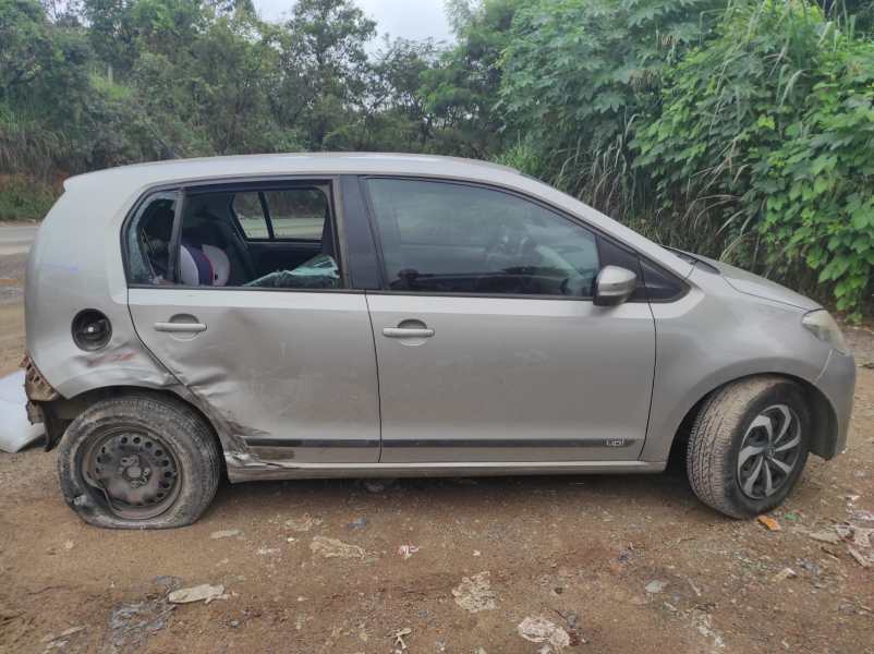 Um carro pequeno e danificado estendido ao lado de uma estrada, com danos visíveis na lateral e um pneu furado, cercado por vegetação.