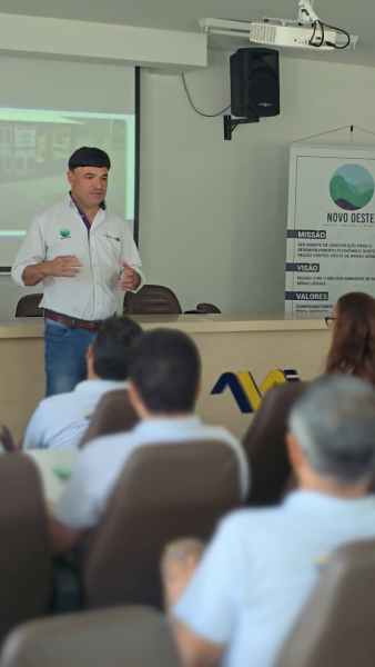 Homem de camisa branca apresentando em uma sala de palestras, com espectadores sentados à frente, projetor ao fundo.