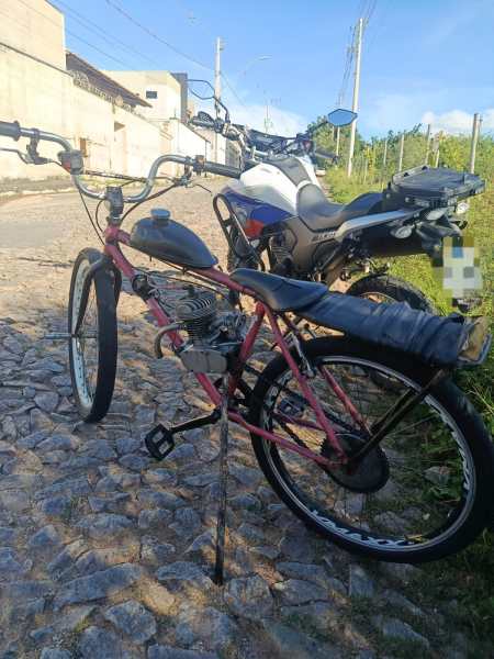 Bicicleta de estilo retrô com motor à combustão, estacionada ao lado de uma moto em uma rua de pedras.
