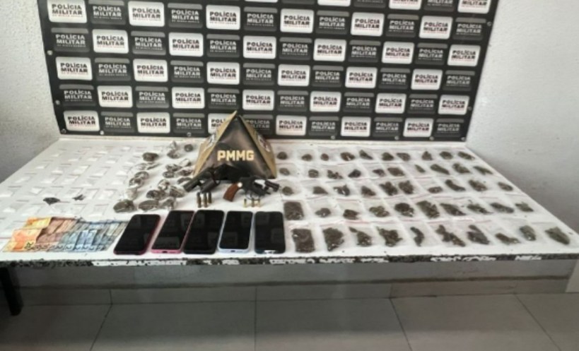 Uma mesa com diversos itens apreendidos, incluindo drogas embrulhadas, quatro celulares, dinheiro e uma placa da Polícia Militar.