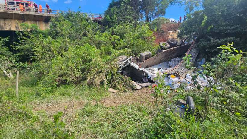 Veículo tombado em um terreno coberto de vegetação, próximo a uma ponte, com equipes de resgate visíveis ao fundo.