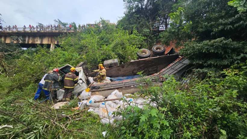 Caminhão tombado em área verde com bombeiros e equipe de resgate trabalhando na cena.