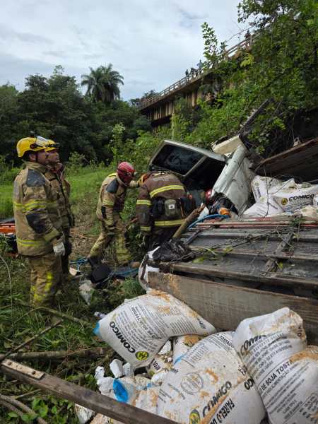 Bombeiros realizando resgate em um local de acidente de carro, com veículo capotado entre vegetação e sacos de cimento.