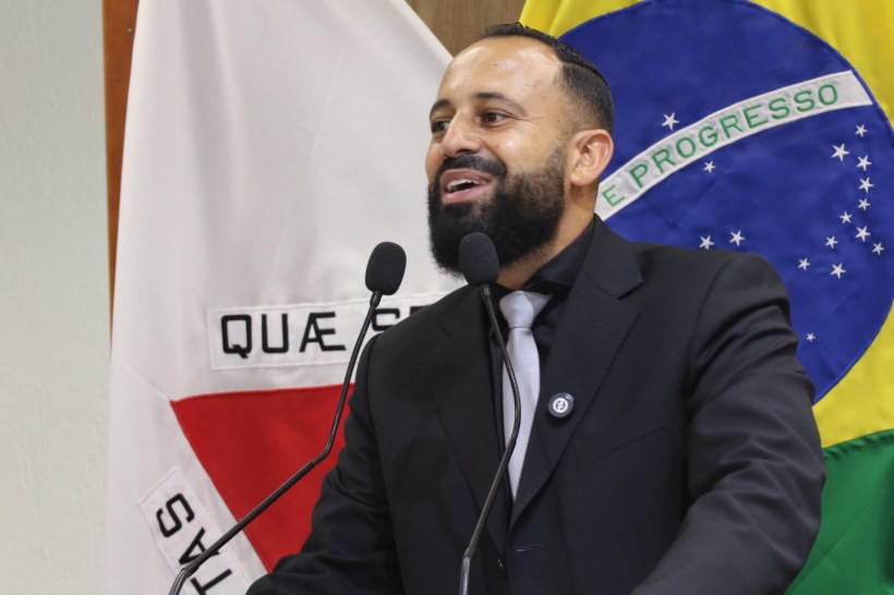 Homem de terno falando em um púlpito, com bandeira do Brasil e outra bandeira ao fundo.