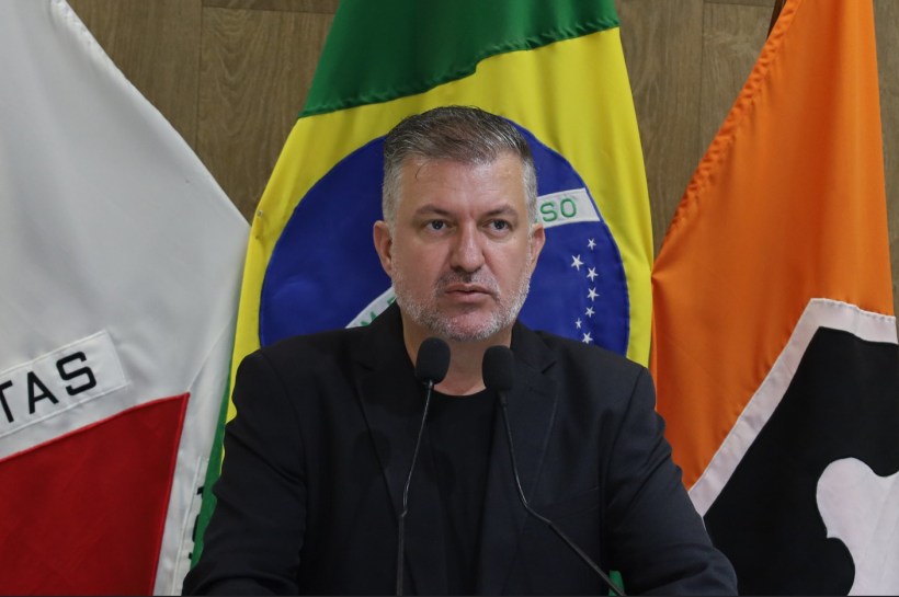Homem de terno preto falando ao microfone em frente a bandeiras do Brasil e outras bandeiras.
