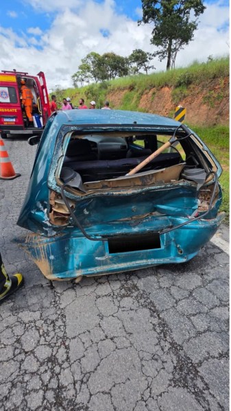 Veículo acidentado com parte traseira danificada em estrada, com bombeiros e equipe de resgate ao fundo.