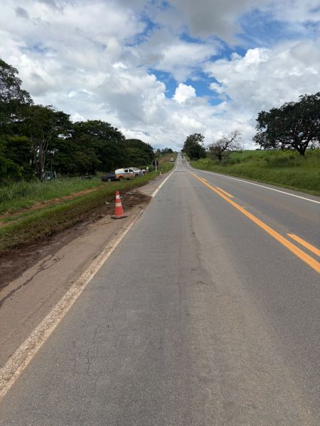 Estrada com árvores ao lado e céu parcialmente nublado, sinalização de obra e cone de segurança visíveis.