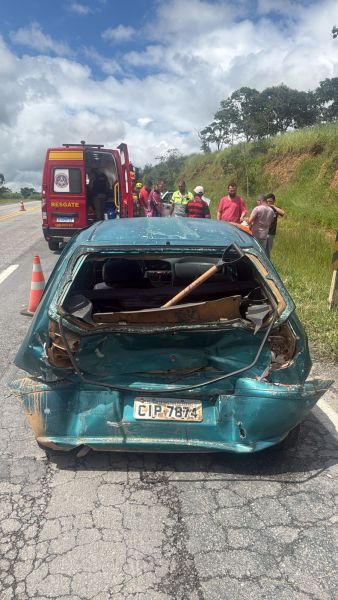Veículo danificado na estrada com parte traseira destruída e equipe de resgate ao fundo.