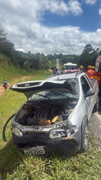 Carro acidentado na estrada, com capô levantado e danos visíveis na frente, enquanto equipe de resgate trabalha ao fundo.