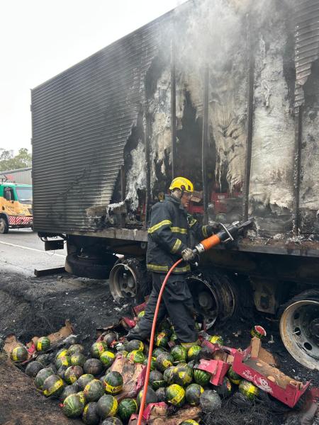 Bombeiro combatendo incêndio em caminhão danificado, com melancias espalhadas no chão.
