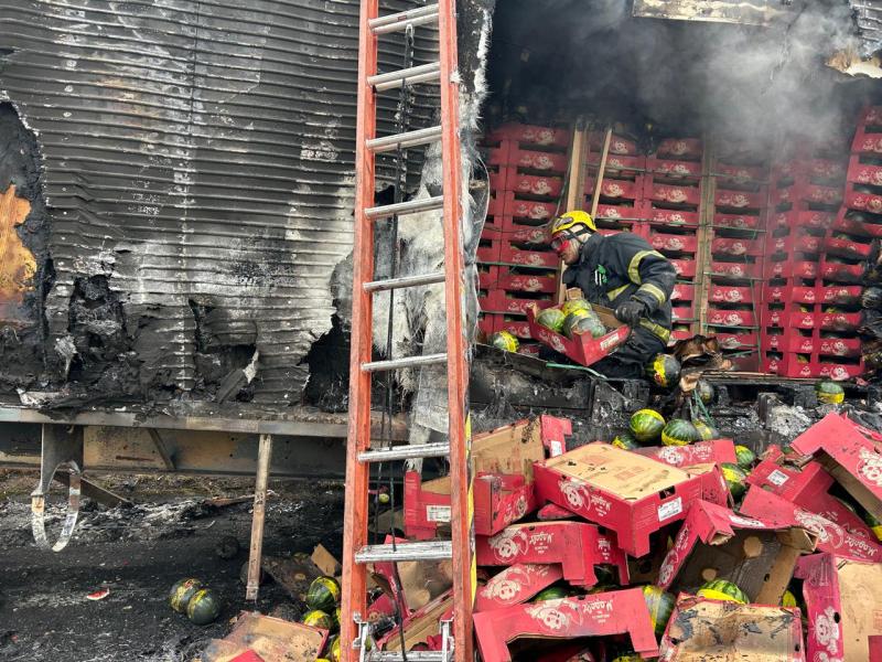 Bombeiro trabalhando em meio a escombros de um incêndio, tirando melancias de caixas danificadas. Ao fundo, uma parede queimada e uma escada.