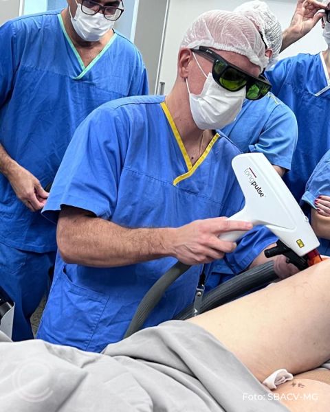 Um médico com máscara e óculos de proteção, vestido com roupas cirúrgicas azuis, utilizando um equipamento de laser em um paciente durante um procedimento médico.