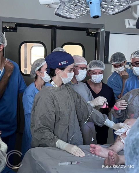Uma equipe médica em ambiente cirúrgico, com profissionais usando máscaras e gorros, assistindo uma cirurgia em um paciente deitado na mesa de operação.
