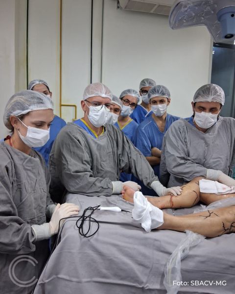 Grupo de profissionais de saúde em uma sala de cirurgia, usando roupas cirúrgicas, toucas e máscaras, observando a execução de um procedimento em um paciente.