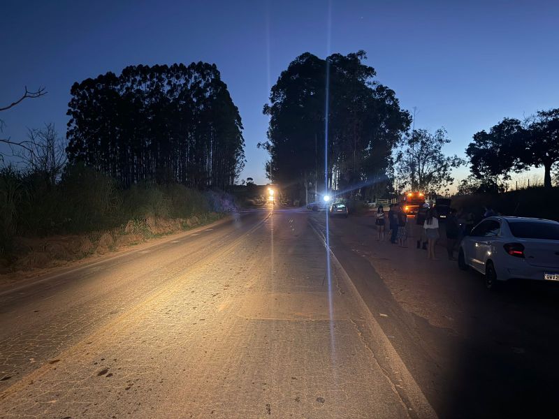 Uma estrada iluminada ao anoitecer, com árvores ao fundo e pessoas em pé ao lado da via.