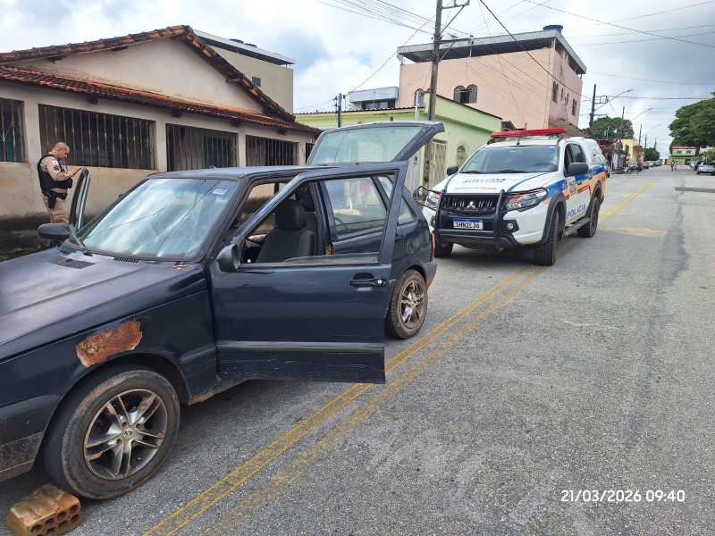Um carro preto estacionado na rua com uma porta aberta e rodas danificadas, ao lado de uma viatura da polícia. Um policial está ao fundo, próximo ao carro.