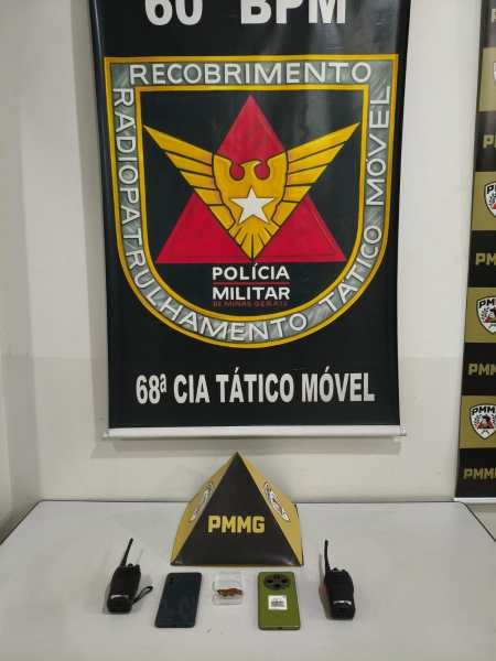 Bandeira da Polícia Militar e equipamentos de comunicação sobre mesa, incluindo rádios e celulares.
