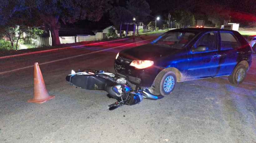 Um carro preto parado na estrada, com uma moto caída ao lado e um cone de sinalização próximo.
