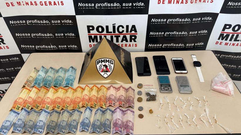 Mesa com dinheiro em espécie, moedas, dispositivos eletrônicos como celulares e relógio, uma pirâmide com o logotipo da Polícia Militar de Minas Gerais e substâncias em sacos plásticos.